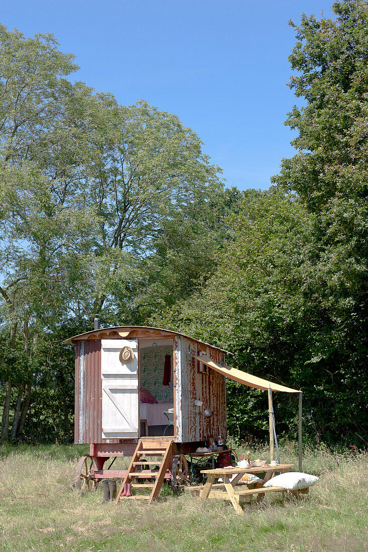 Eine Schäferhütte auf einer Sommerwiese mit einem Zeltdach für eine provisorische Essküche und einer offenen Tür mit Stufen zum Innenraum