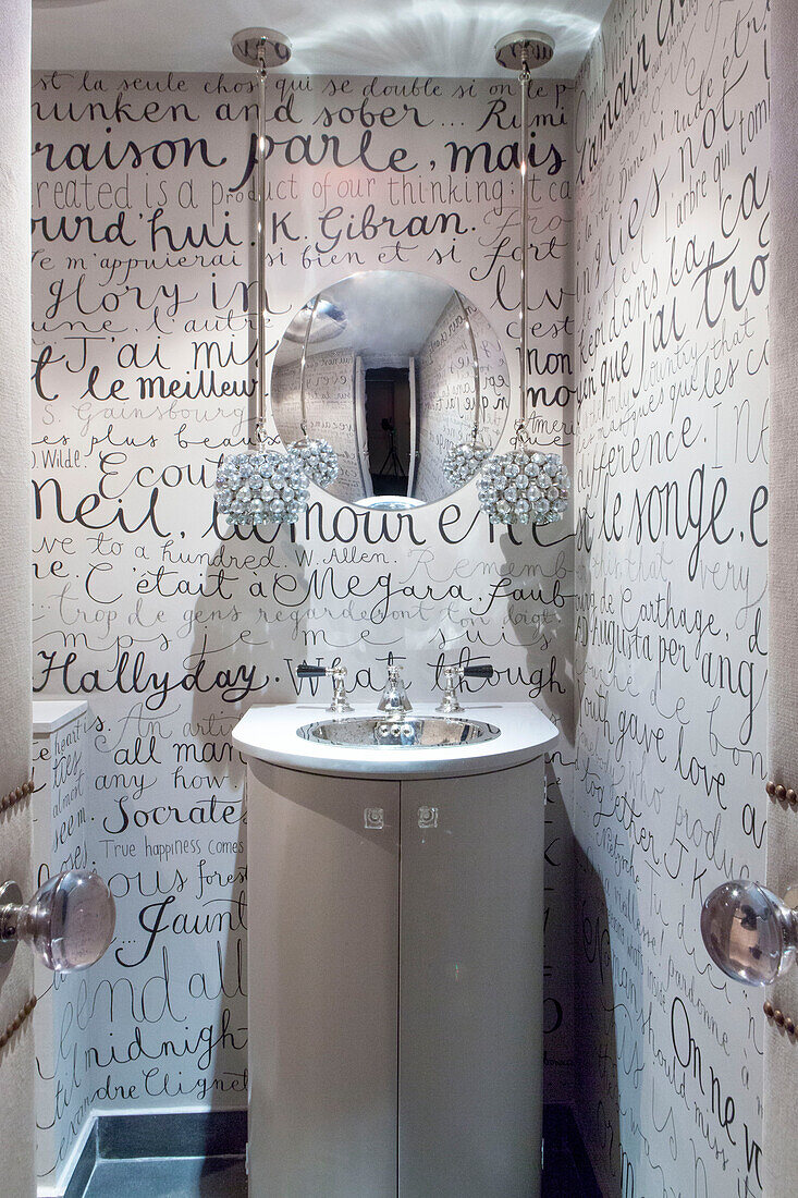 Runder Spiegel mit Lampenschirmen aus geschliffenem Glas und klein geschriebener französischer Texttapete mit Waschbecken in einem modernen Haus in London, England, UK