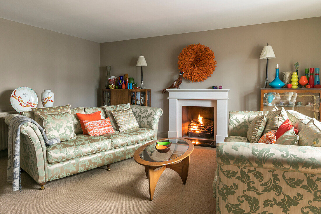 Zwei blattgemusterte Sofas mit beleuchtetem Feuer im Wohnzimmer in Farnham, Surrey, UK