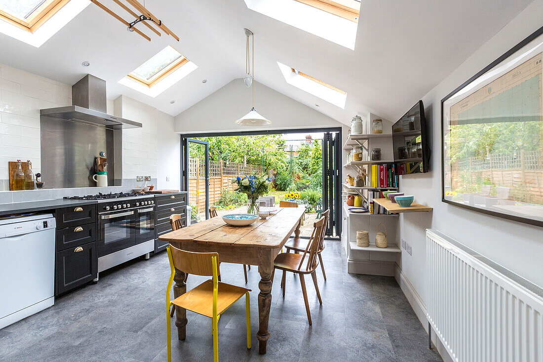 Holztisch und -stühle mit Oberlicht und offenen Türen zur Gartenterrasse in der Küche von Reading, Berkshire, England UK