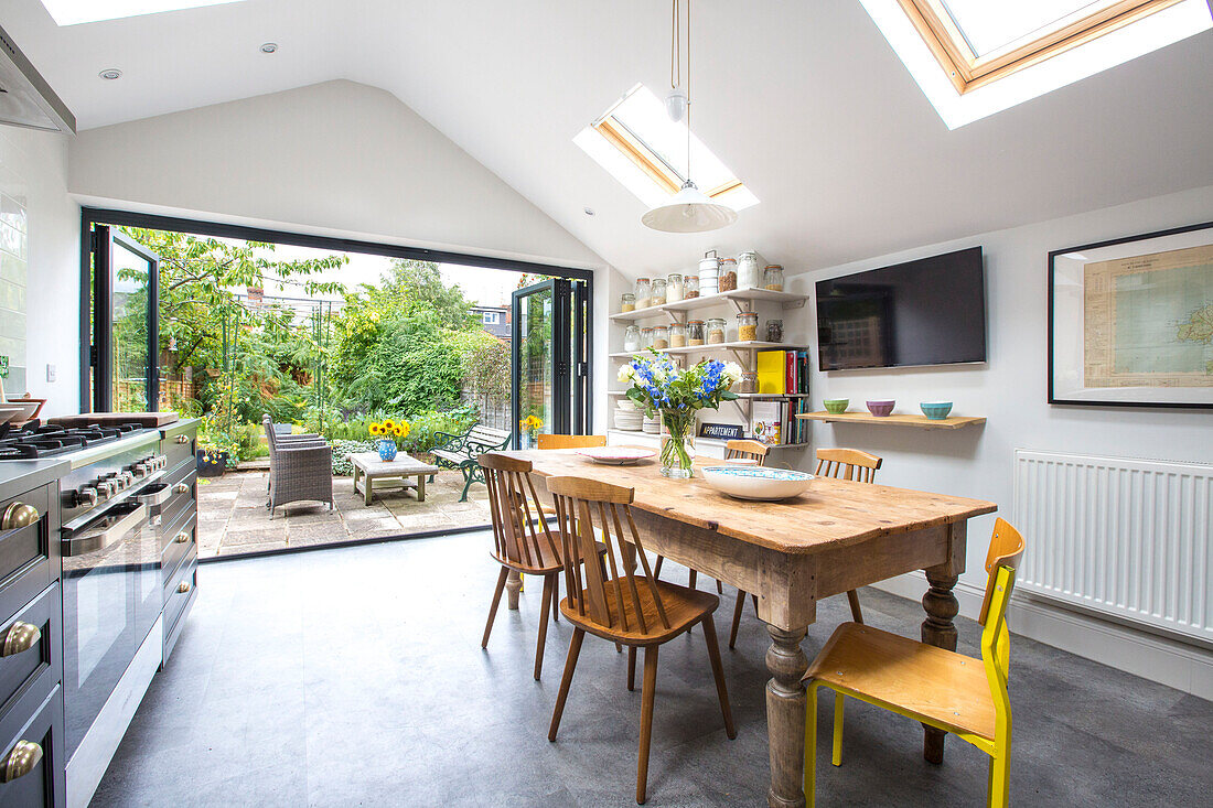 Holztisch und Stühle unter Dachfenstern mit offenen Türen zur Terrasse in der Küche von Reading, Berkshire, England UK