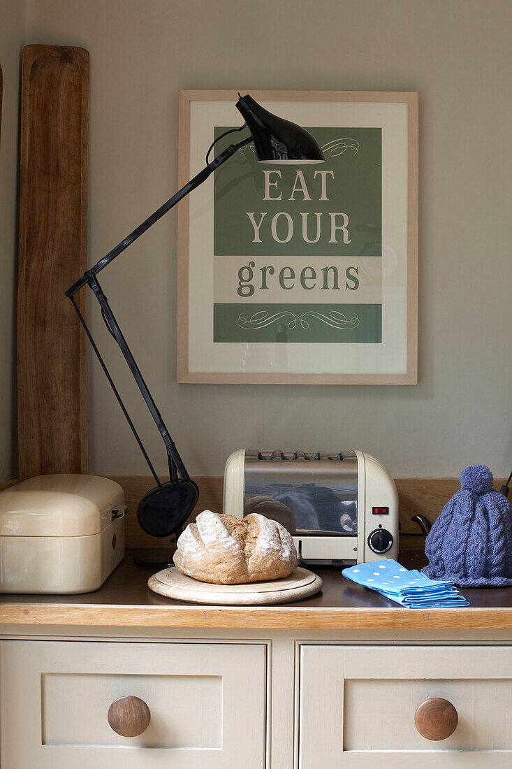 Frisches Brot in einer Küche mit Winkellampe und einem Poster mit der Aufschrift 'Eat Your Greens' (Iss dein Grün)