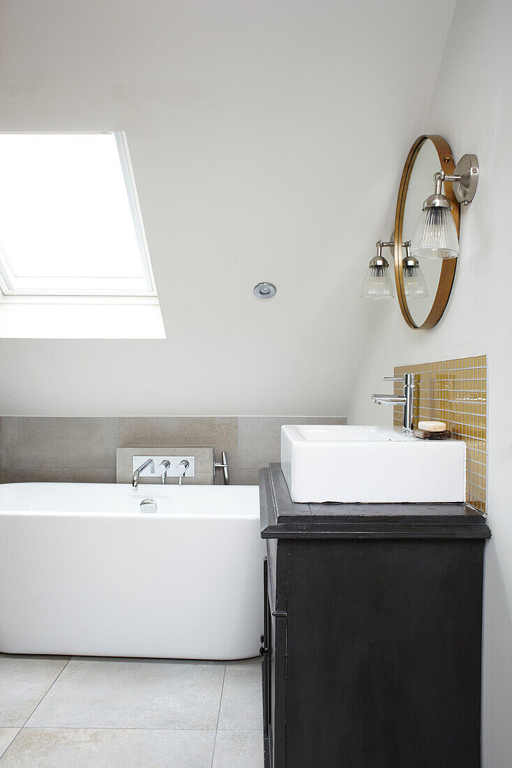 Circular mirror above wash basin with freestanding bath below skylight in attic bathroom Brighton home East Sussex UK