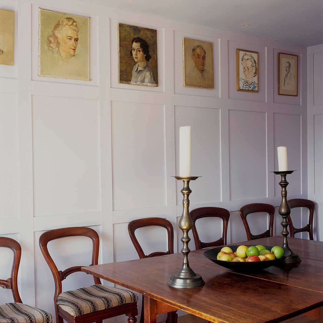 Dining room with oak dining table and chairs set with pewter candlesticks and a fruit bowl full of apples lilac painted wood panels and portraits on the wall