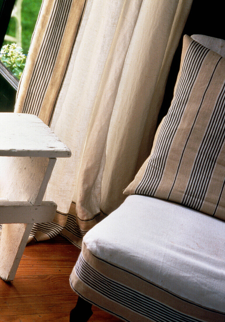 Detail eines mit Leinenstoff bezogenen Stuhls neben einem Fenster mit Leinenvorhang
