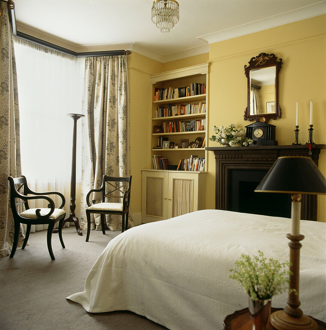 In blassem Gelb gestrichenes Schlafzimmer mit eingebautem Bücherregal und Erkerfenster mit zwei antiken Sesseln und weißen Blumenarrangements
