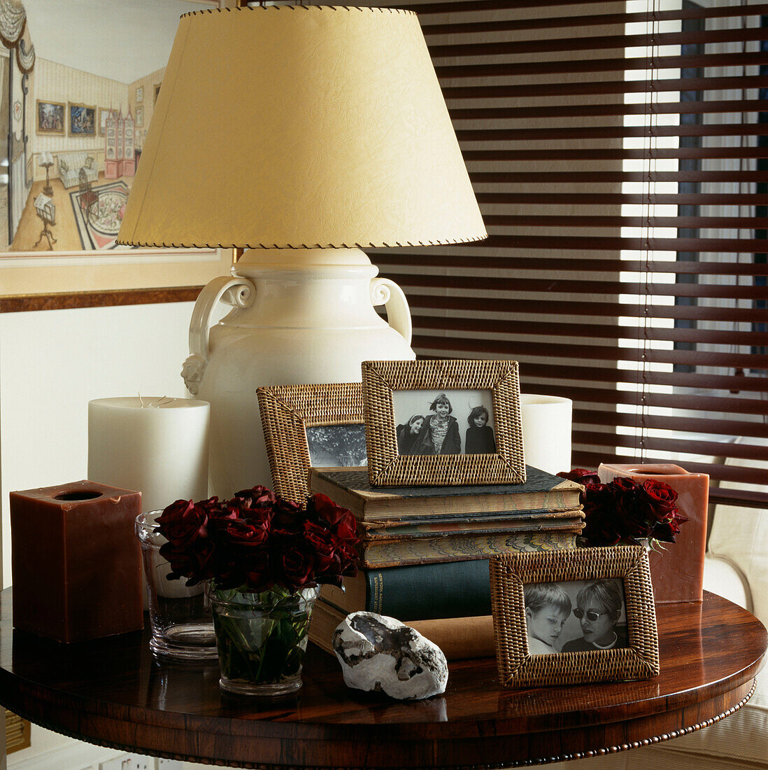 Corner of room with Venetian blind and small table with candles and table lamp and vases of roses and family photos and books