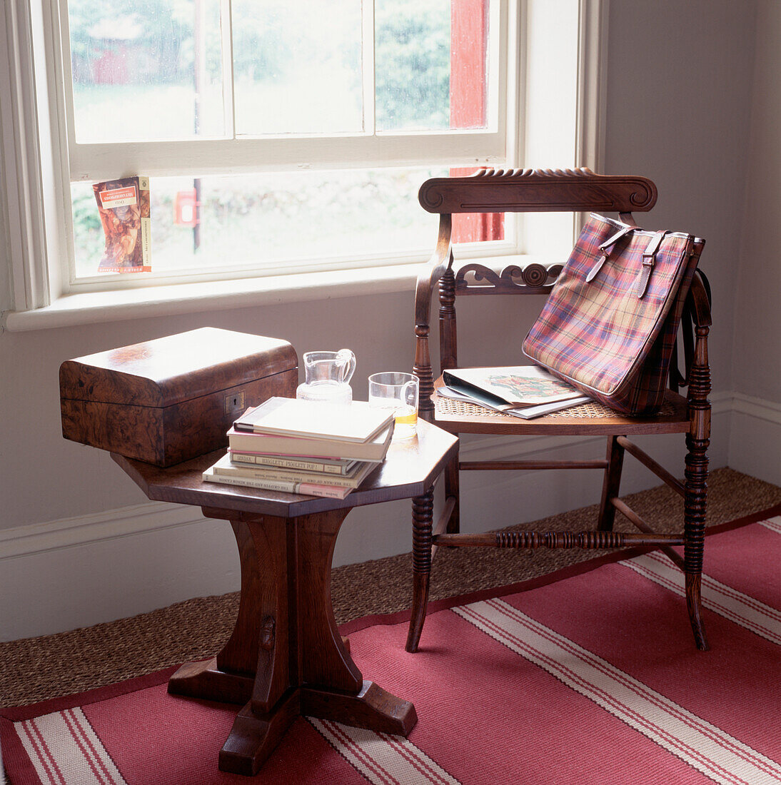 Eichenstuhl mit karierter Tasche neben einem Beistelltisch mit Holzkiste und aufgestütztem Wasserglasfenster mit einem Buch im Hintergrund