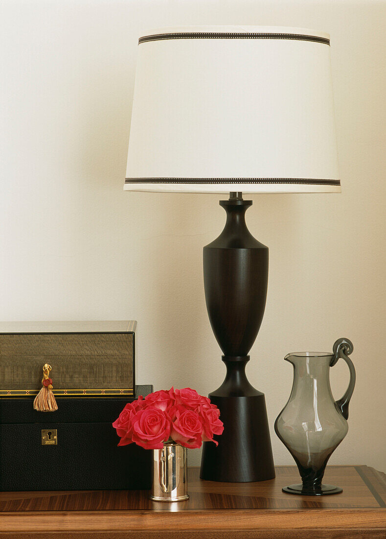 Elegante schwarze Tischlampe mit weißem Schirm auf Tischplatte mit Glaskanne und silberner Vase mit Rosen gefüllt