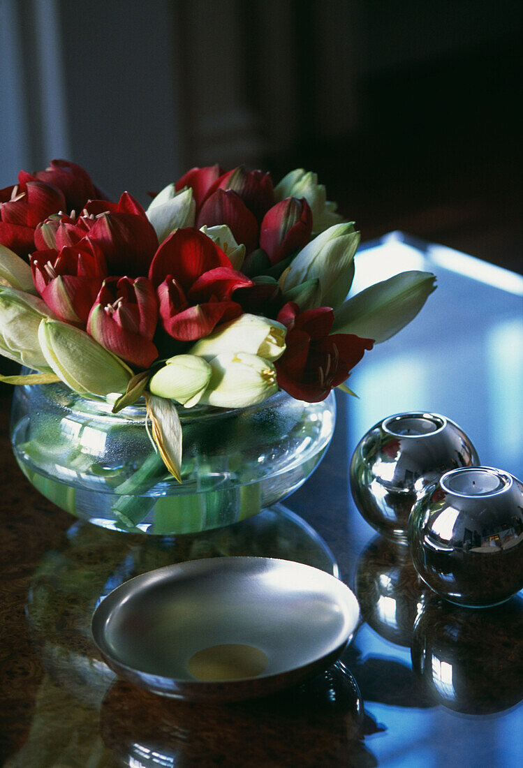 Mit Tulpen gefüllte Glasschale auf einer glänzenden Tischplatte mit Kerzenhaltern und Schale aus Metall