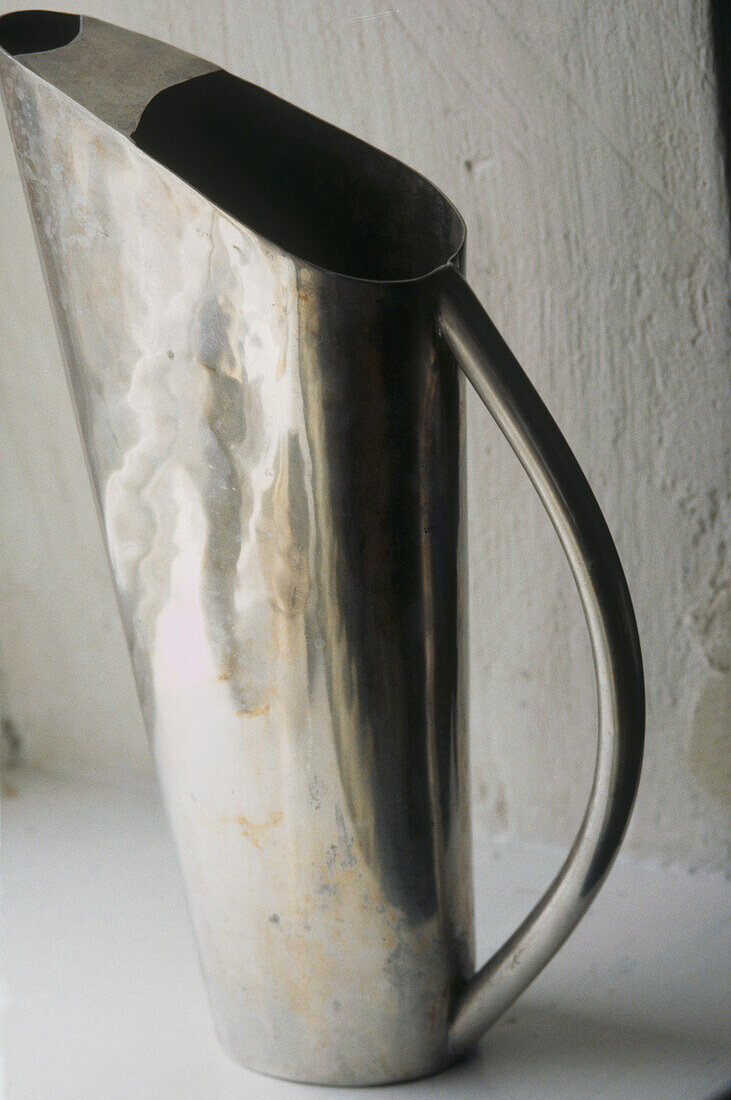 Close up of an unusual shaped silver coloured jug against white plaster wall