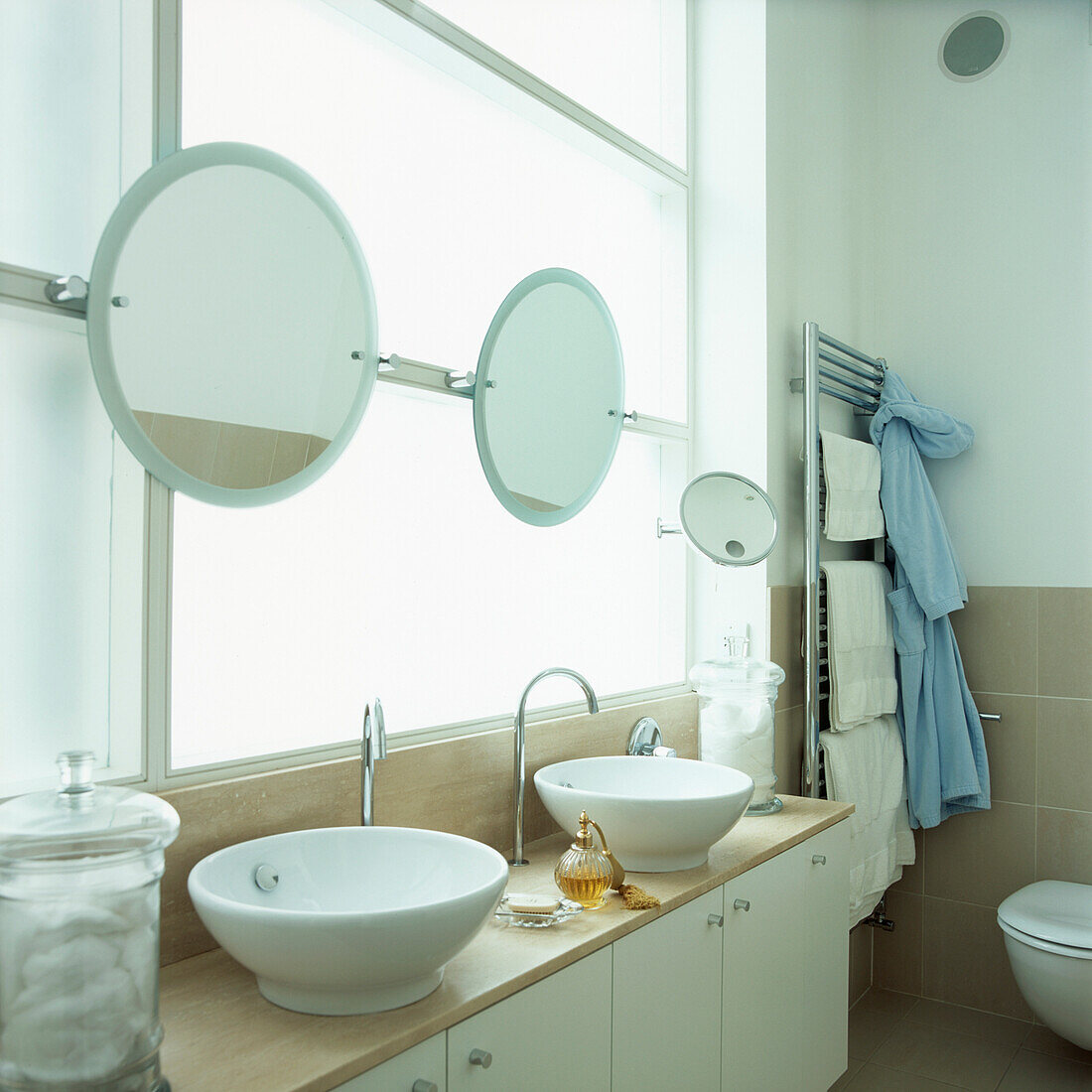 Modernes weißes Badezimmer mit zwei Waschbecken, Spiegeln, Ablageschränken und einem großen Fenster aus Milchglas