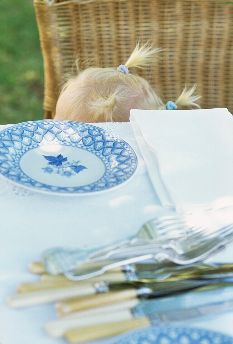 Gartentisch, darunter Kopf eines kleinen Mädchens mit blonden Haaren und kleinen Zöpfchen