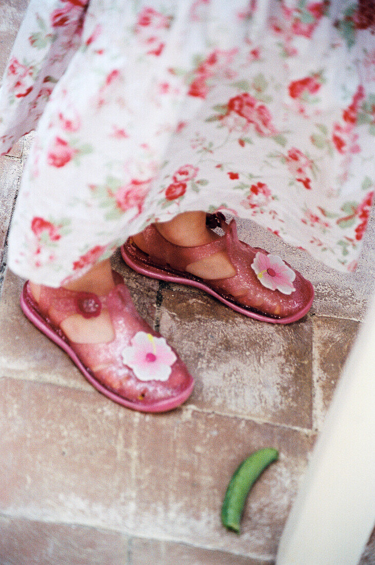 Detail of young girl's floral skirt and jelly bean sandals with broad bean on floor