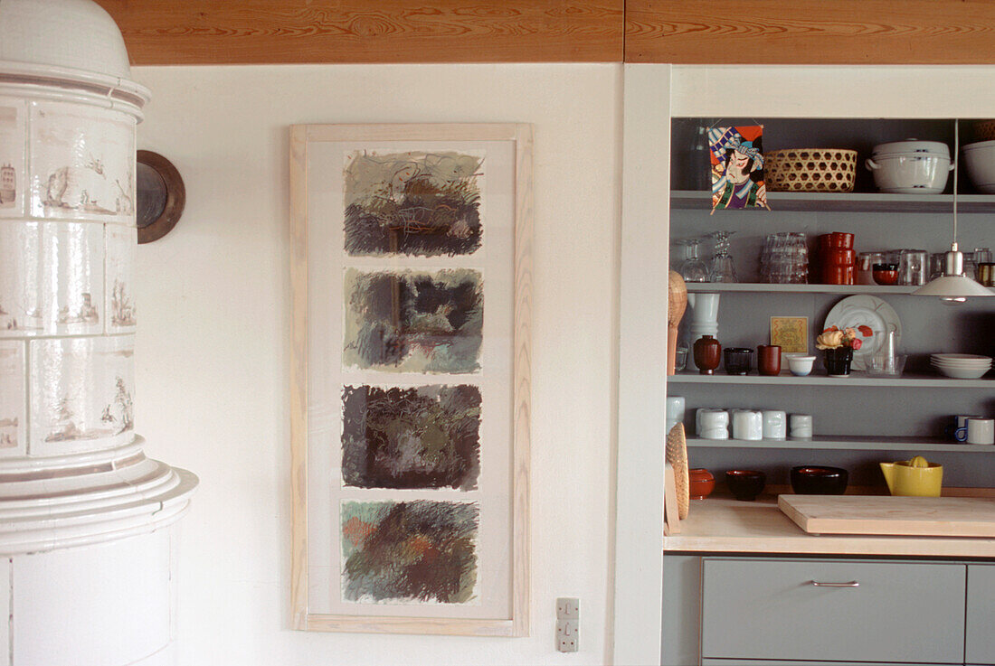 Weißer Kachelofen in einer Küche mit einer grau gestrichenen Kommode, auf der Küchen- und Tafelgeschirr aufbewahrt wird