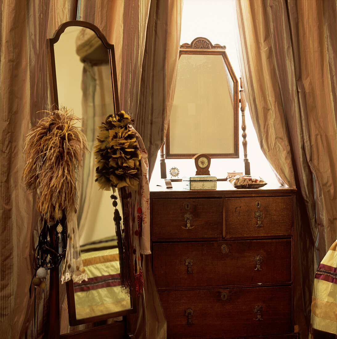 Bedroom detail with Cheval mirror and feather trimmings next to antique chest of drawers with uncut silk ribbon curtains