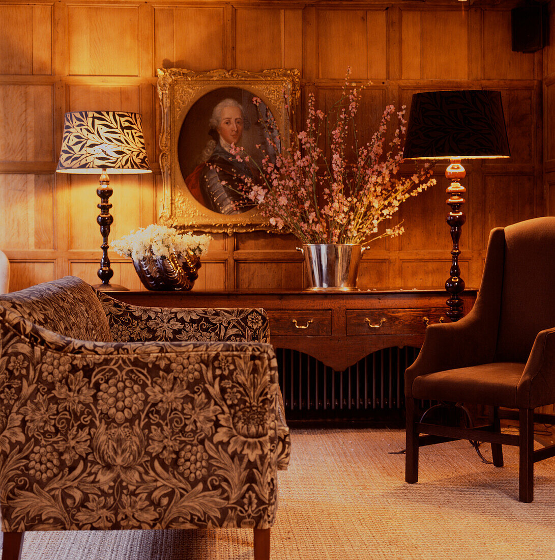 Floral print sofa and armchair in wood panelled lounge with large bouquet and wooden lamps illuminating the room
