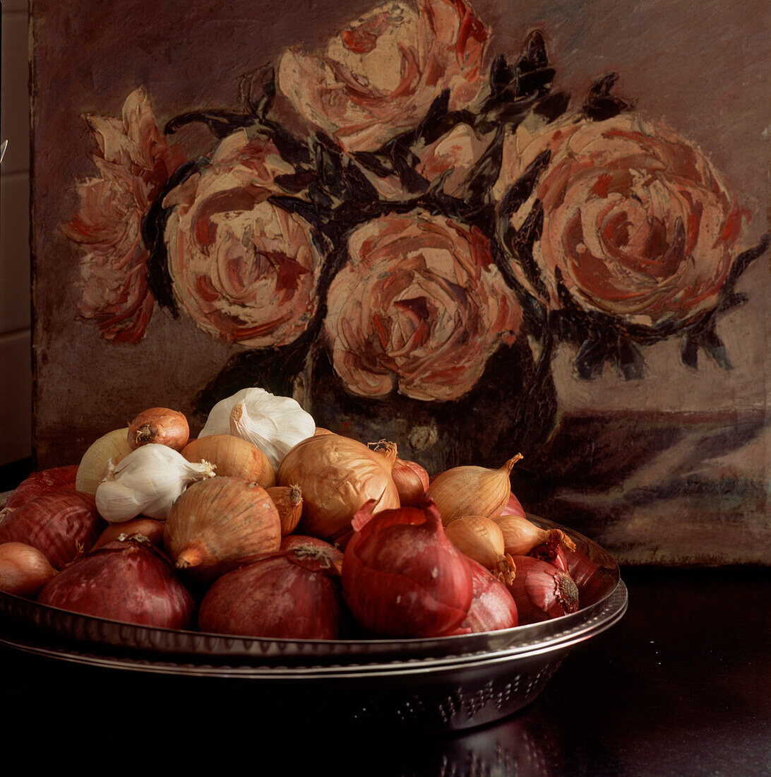 Still life with bowl of onions and garlic on tabletop with a floral oil painting in background