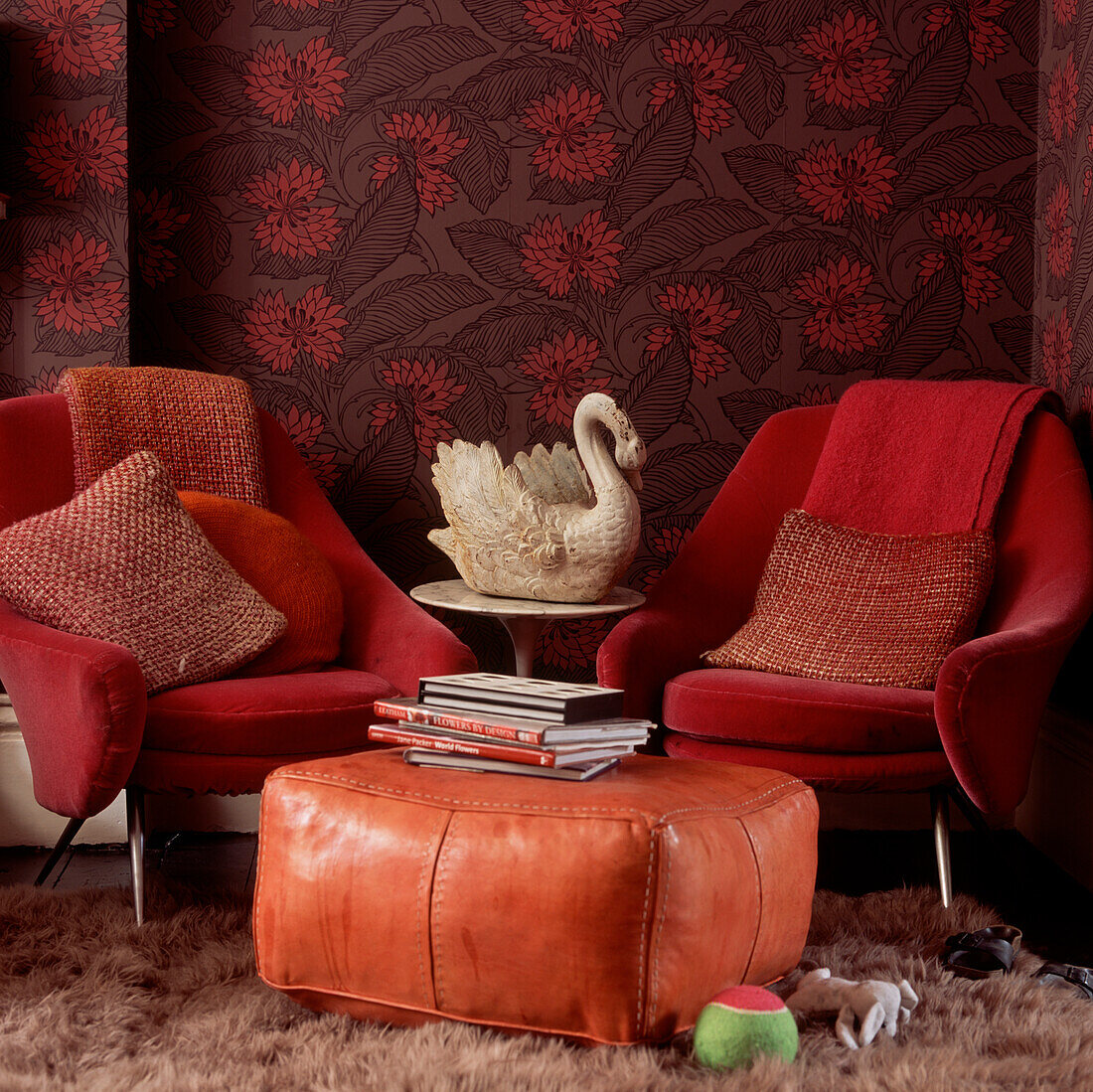 Upholstered armchairs and leather floor cushion on shag pile rug with dark floral wallpaper and a swan vase