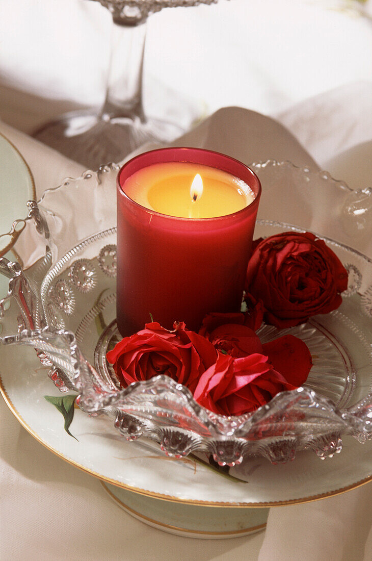 Glühende Kerze auf einer verzierten Glasschale mit roten Rosen auf einem Tisch