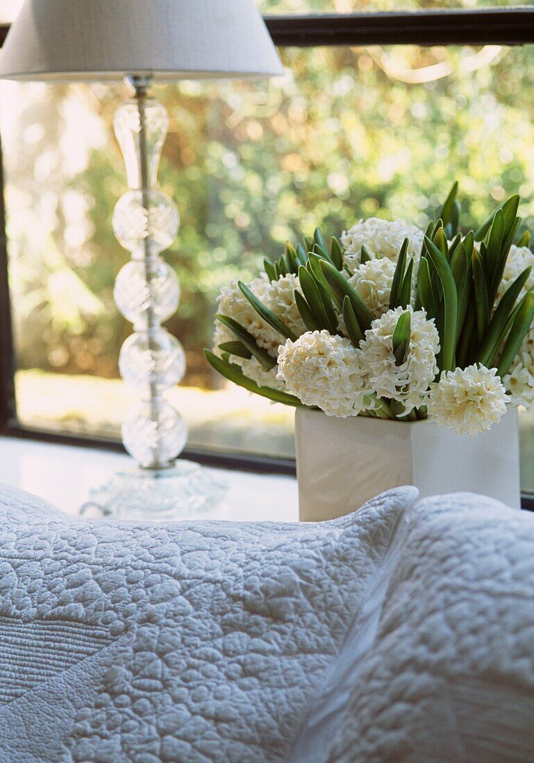 Nahaufnahme einer weiß bestickten Tagesdecke mit einer Vase mit weißen Hyazinthen und einem gläsernen Lampenständer an einem Fenster
