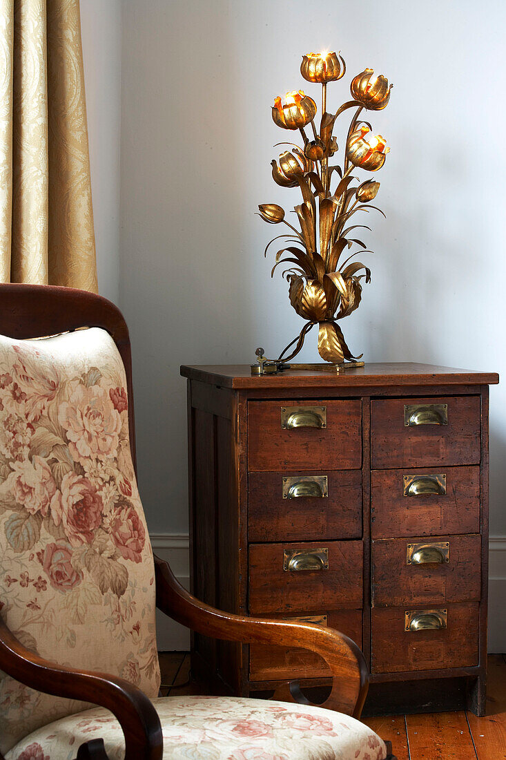 Ornate light on antique wooden drawers with armchair in London townhouse, UK