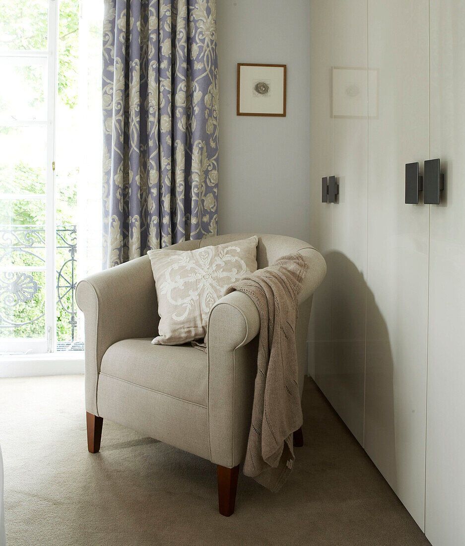 In sanften Pastellfarben dekorierte Schlafzimmerecke mit gepolstertem Sessel und gemusterten lila Vorhängen