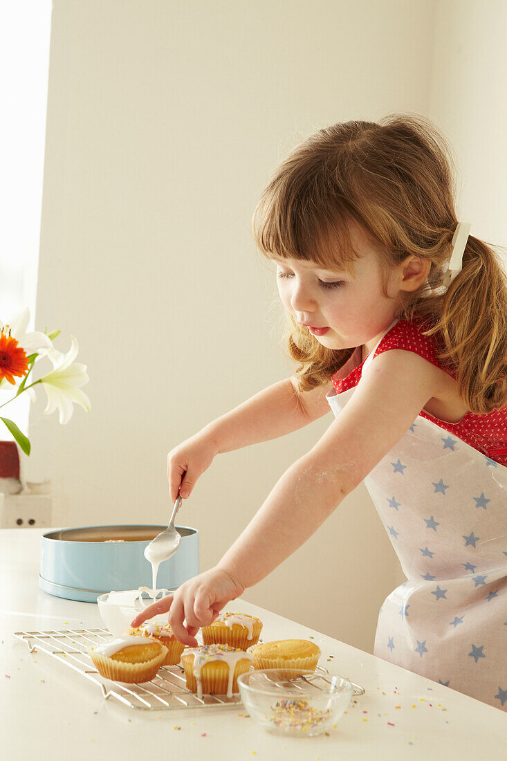 Drei Jahre altes Mädchen steht und glasiert Fairy cakes
