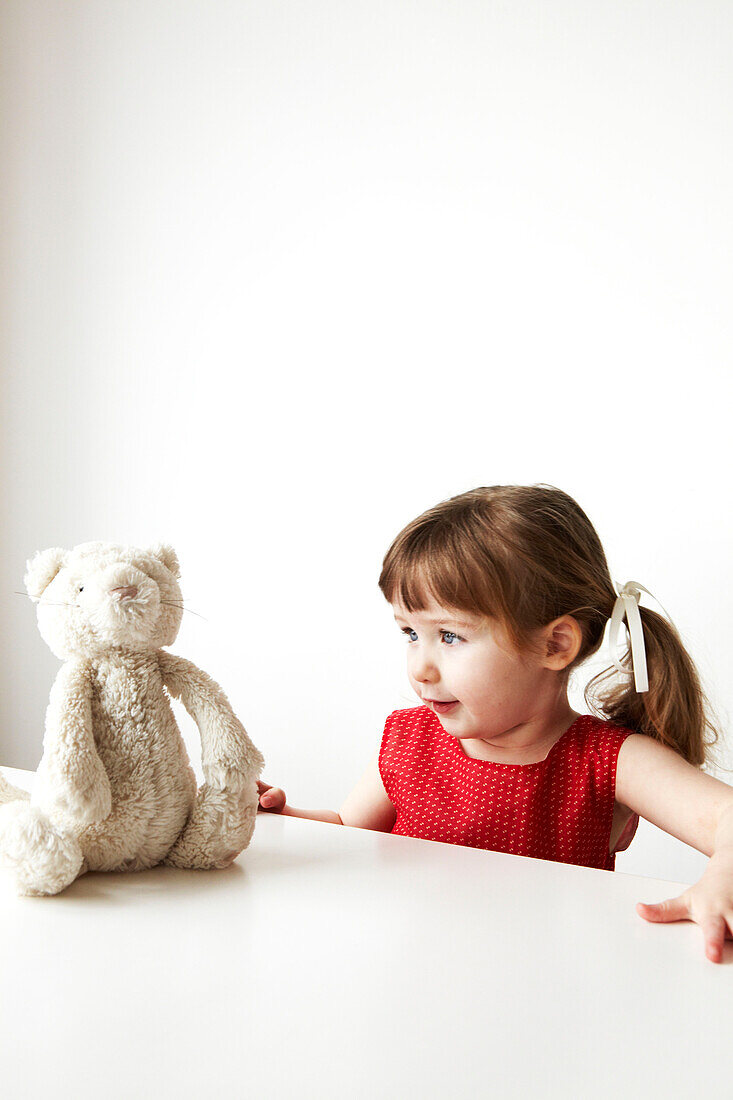 Junges Mädchen in rotem Kleid sitzt da und schaut ihren Teddybär an