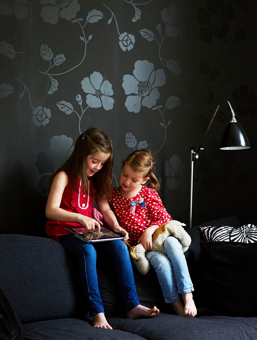 Two sisters sitting on sofa with floral patterned wallpaper in London townhouse, England, UK