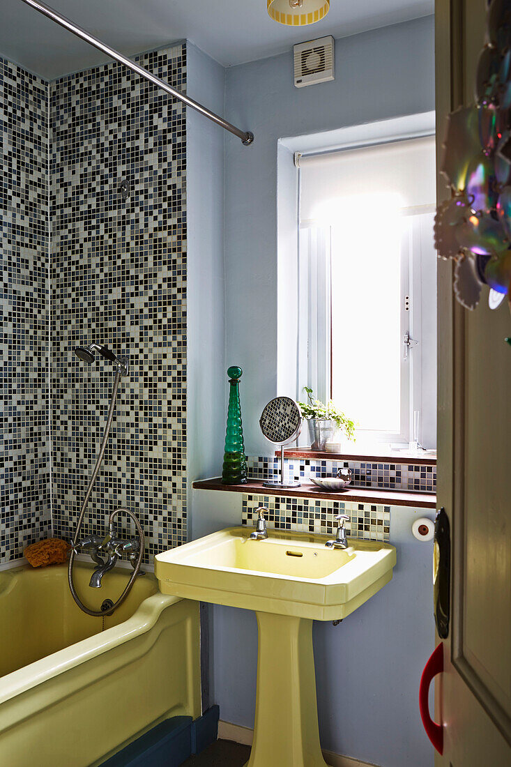 Yellow bath and sink at window of mosaic tiled bathroom of Hackney home, East London, UK