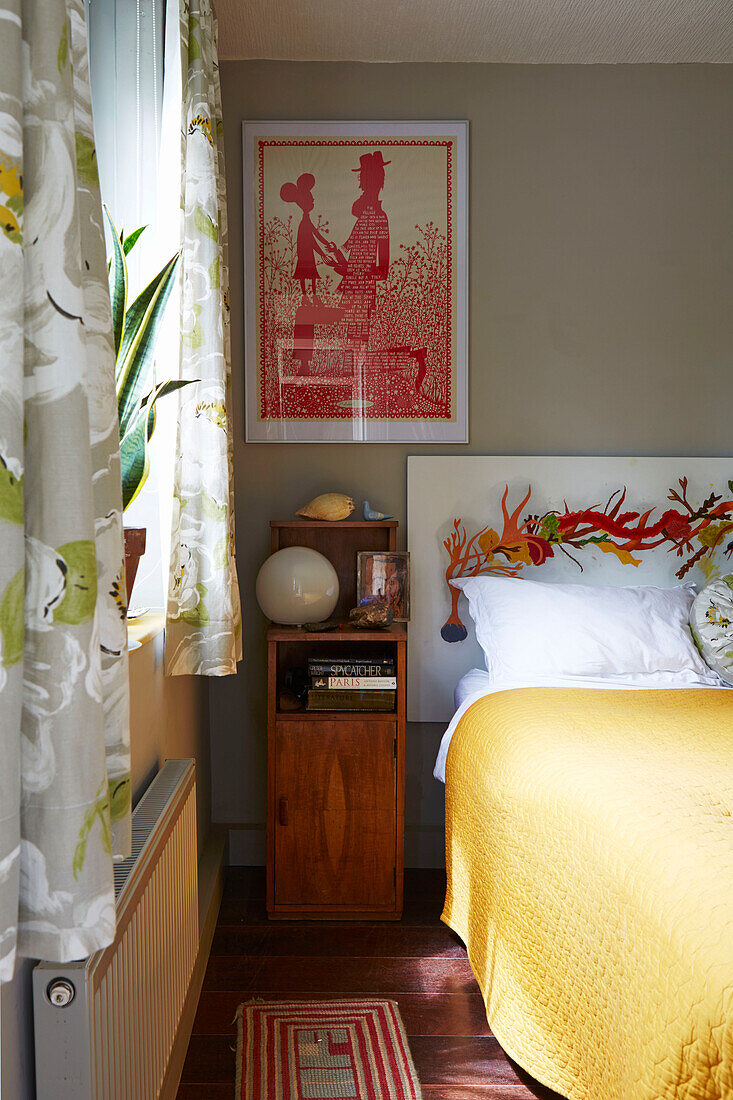 Wooden bedside cabinet with yellow bedspread at window in Hackney bedroom, East London, UK