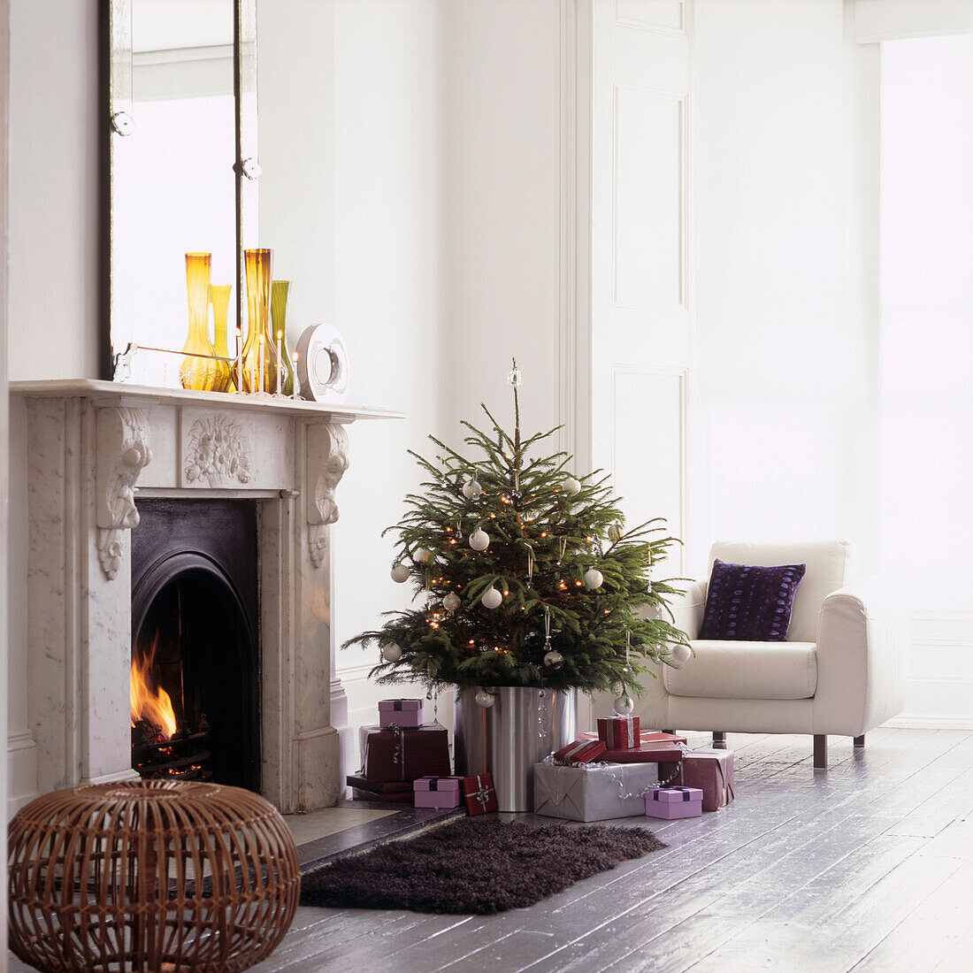 Sonnendurchflutetes weißes Wohnzimmer mit Weihnachtsbaum und Geschenken vor beleuchtetem Feuer