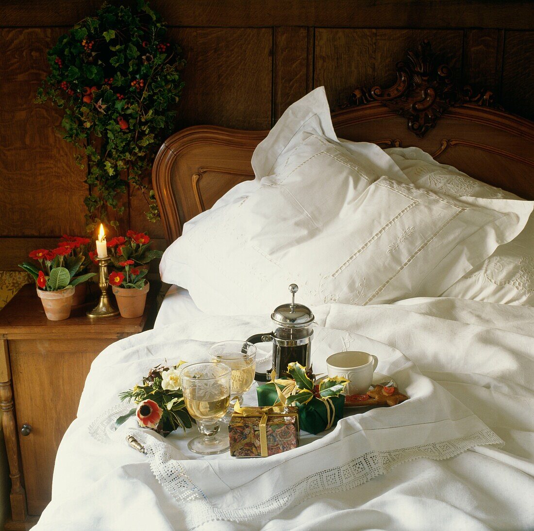 Frühstückstablett mit Kaffeemaschine auf weißer Bettwäsche mit brennenden Kerzen und Zimmerpflanzen auf dem Nachttisch