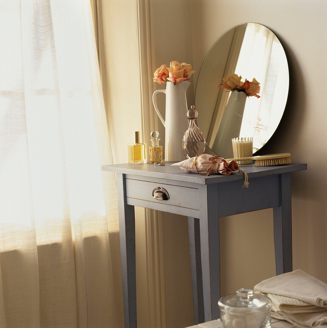 Vase mit Blumen und kreisförmigem Spiegel auf bemaltem Nachttisch an einem mit Vorhängen versehenen, sonnenbeschienenen Fenster