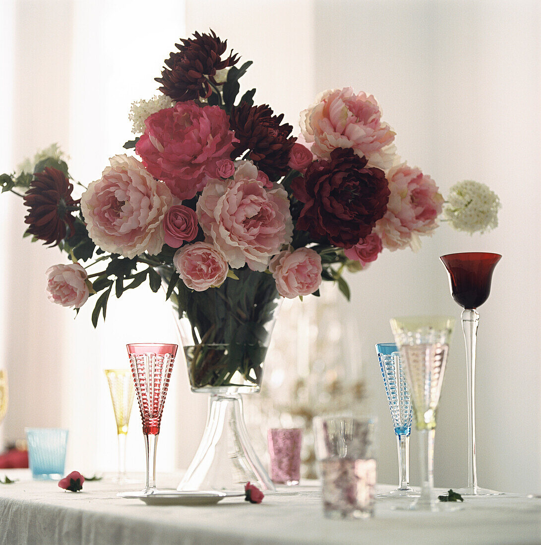 Large bouquet of Peonies in glass vase on a table top with decorative coloured wine glasses