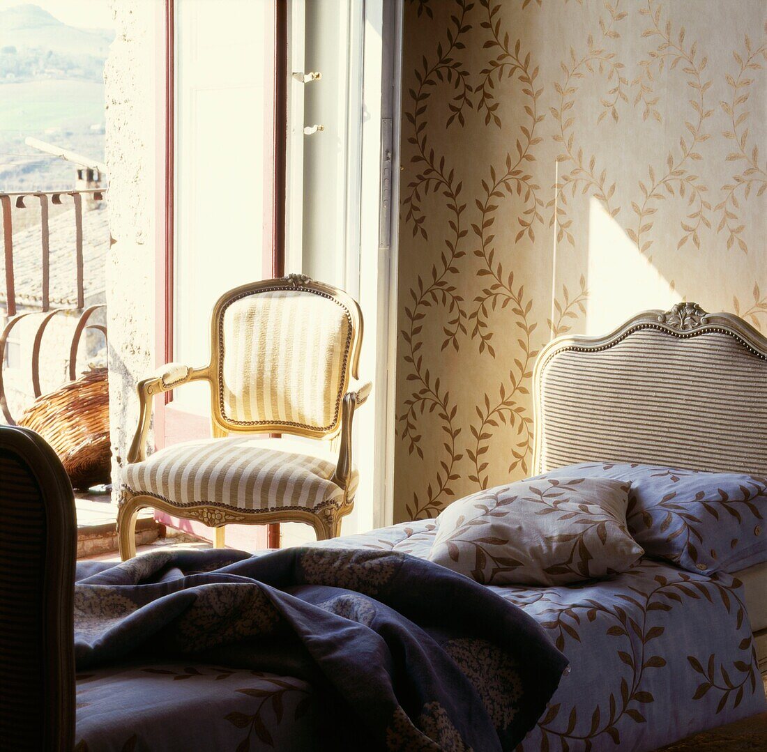 Sessel vor offener Tür in einem sonnendurchfluteten Schlafzimmer mit abgestimmter Tapete und Bettwäsche
