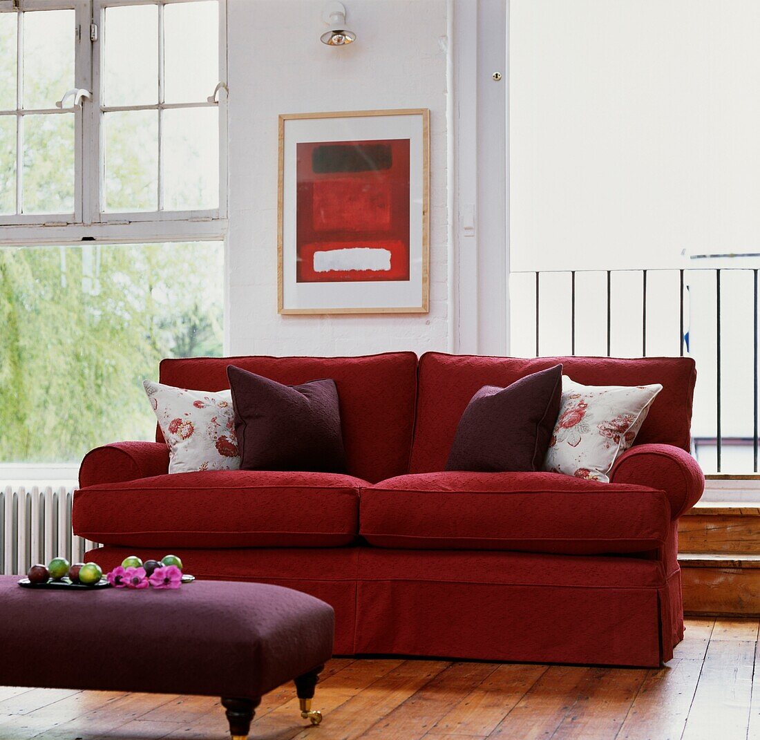 Rotes Sofa mit lilafarbenen Kissen und Fußhocker unter einem modernen Kunstwerk zwischen Schiebefenster und offener Balkontür