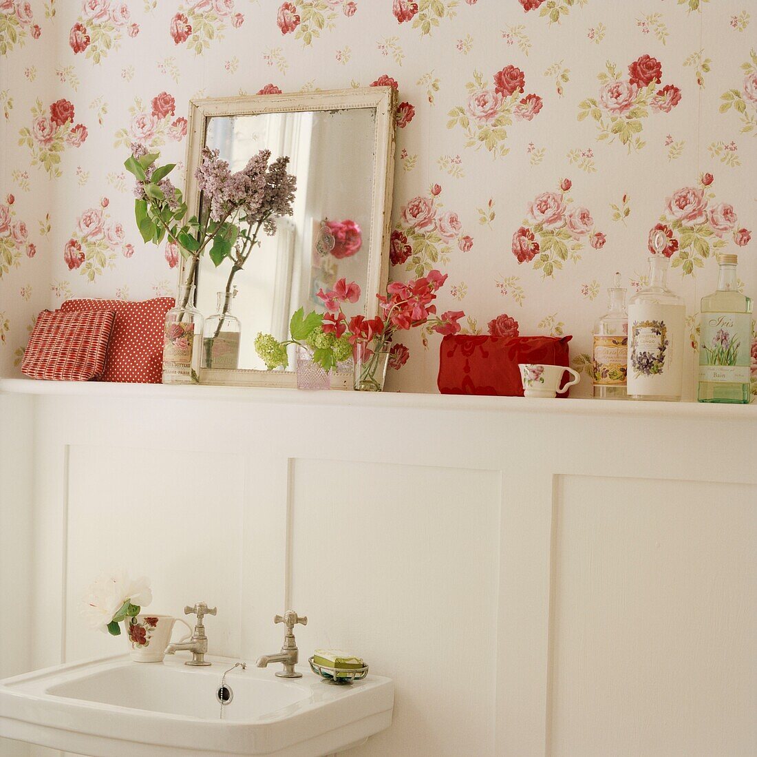 Cut flowers and toiletries on bathroom shelf with floral patterned wallpaper and mirror