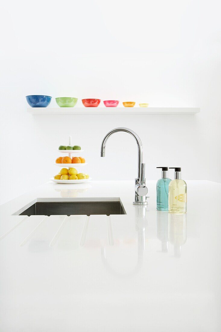 Kitchen sink with chrome mixer tap built into white worktop with bottles of soap a shelf of colourful bowls and a cakestand with citrus fruit