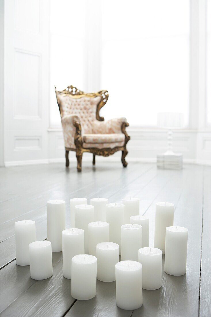 18 unbeleuchtete Kirchenkerzen auf gestrichenen Dielen in einem minimalistisch eingerichteten, ganz in Weiß gehaltenen Raum mit Erkerfenster und antikem Sessel