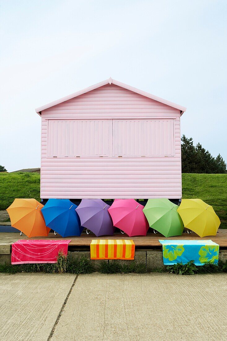 Rosa Strandhütte auf einer Grasbank mit mehrfarbigen Sonnenschirmen in einer Reihe und Strandtüchern