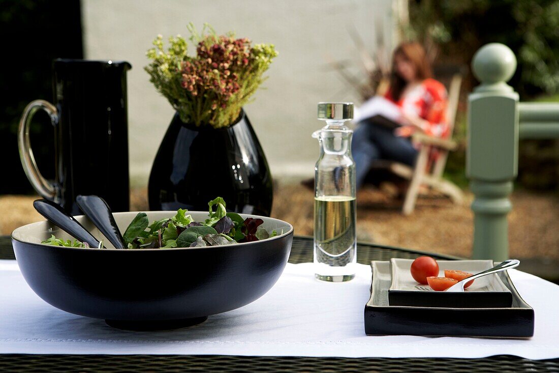 Gedeckter Gartentisch mit Salat und Geschirr, im Hintergrund sitzt eine Frau