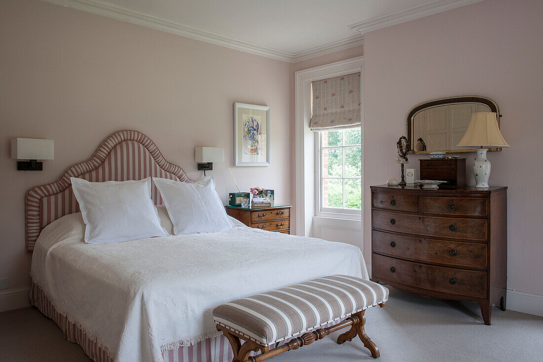 Neutrally decorated double bedroom with upholstered bedhead and bench with antique mahogany chest of drawers
