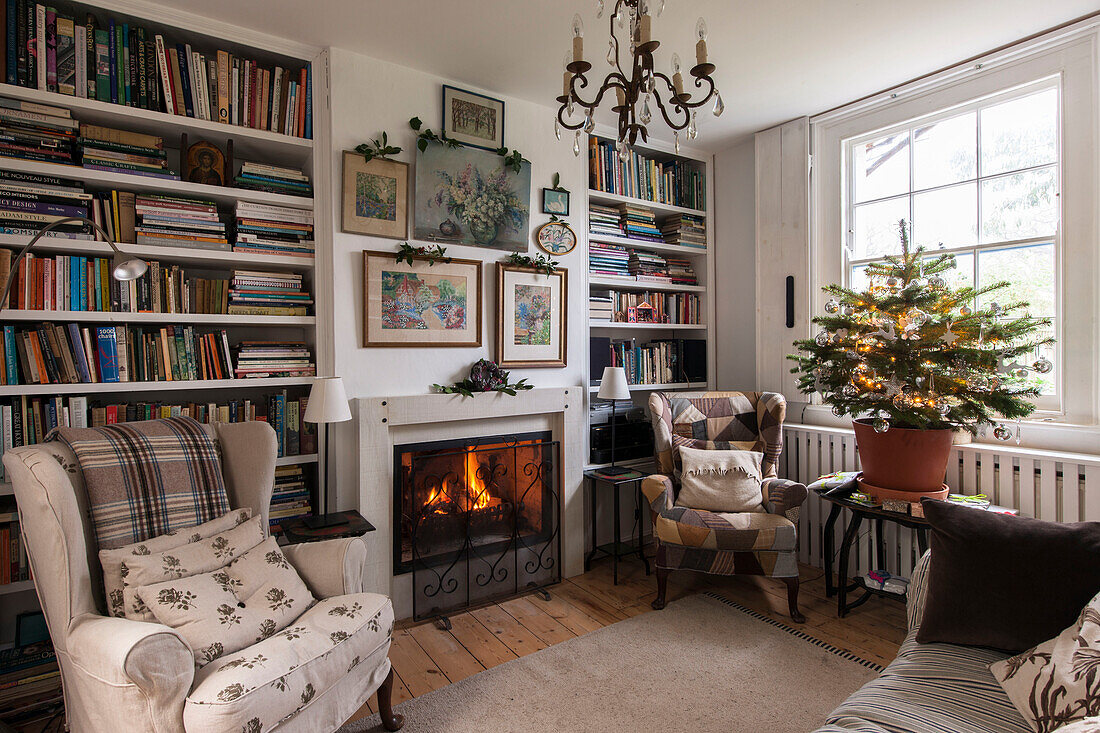 Sessel vor beleuchtetem Kamin mit Bücherregalen und Weihnachtsbaum in einem Londoner Haus in England UK