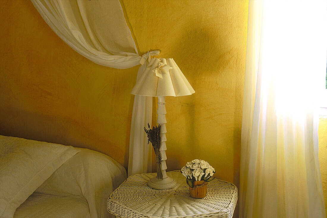 Schlafzimmer mit Nachttisch aus Weidengeflecht und weißen Vorhängen
