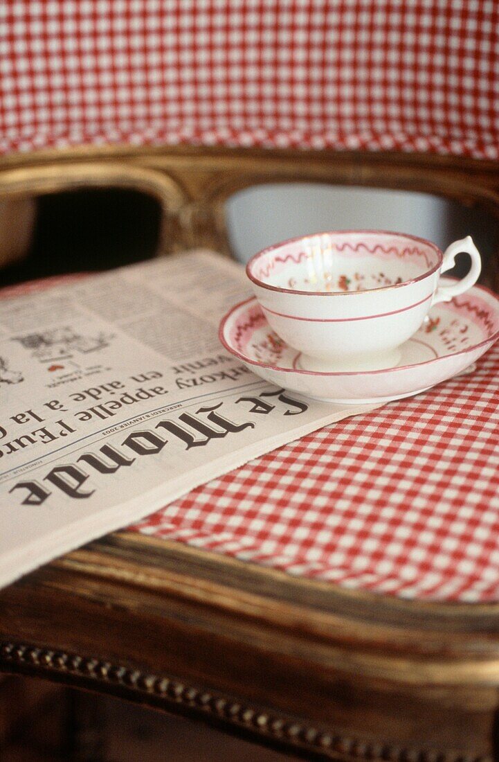 Detail of cup and saucer on a gingham vintage chair with a newspaper