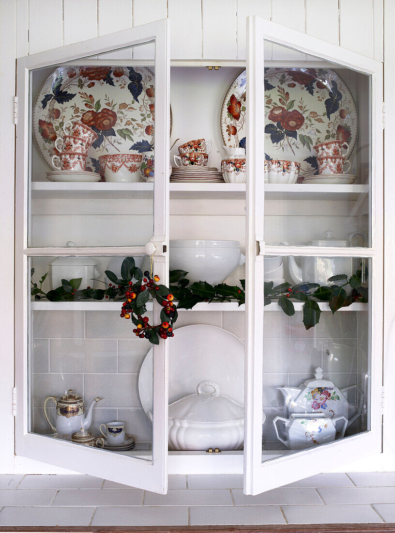 Glass cabinet with open doors showing tiled work surface and wall filled with vintage dinnerware and decorated with a holly garland and wreath