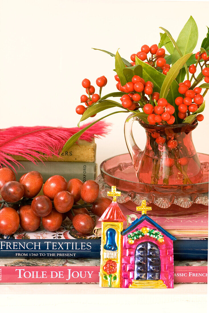 Stilleben mit Textilbüchern, einem mit roten Beeren und grünen Blättern gefüllten Krug aus Preiselbeerglas und einer Kapelle aus Pressblech
