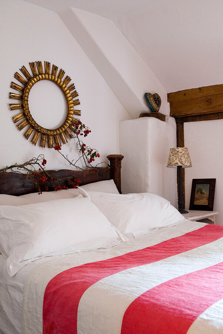 Schlafzimmer mit rot-weiß gestreifter Tagesdecke und einem runden vergoldeten Rahmen, der über Zweigen mit roten Beeren am Kopfende des Bettes hängt